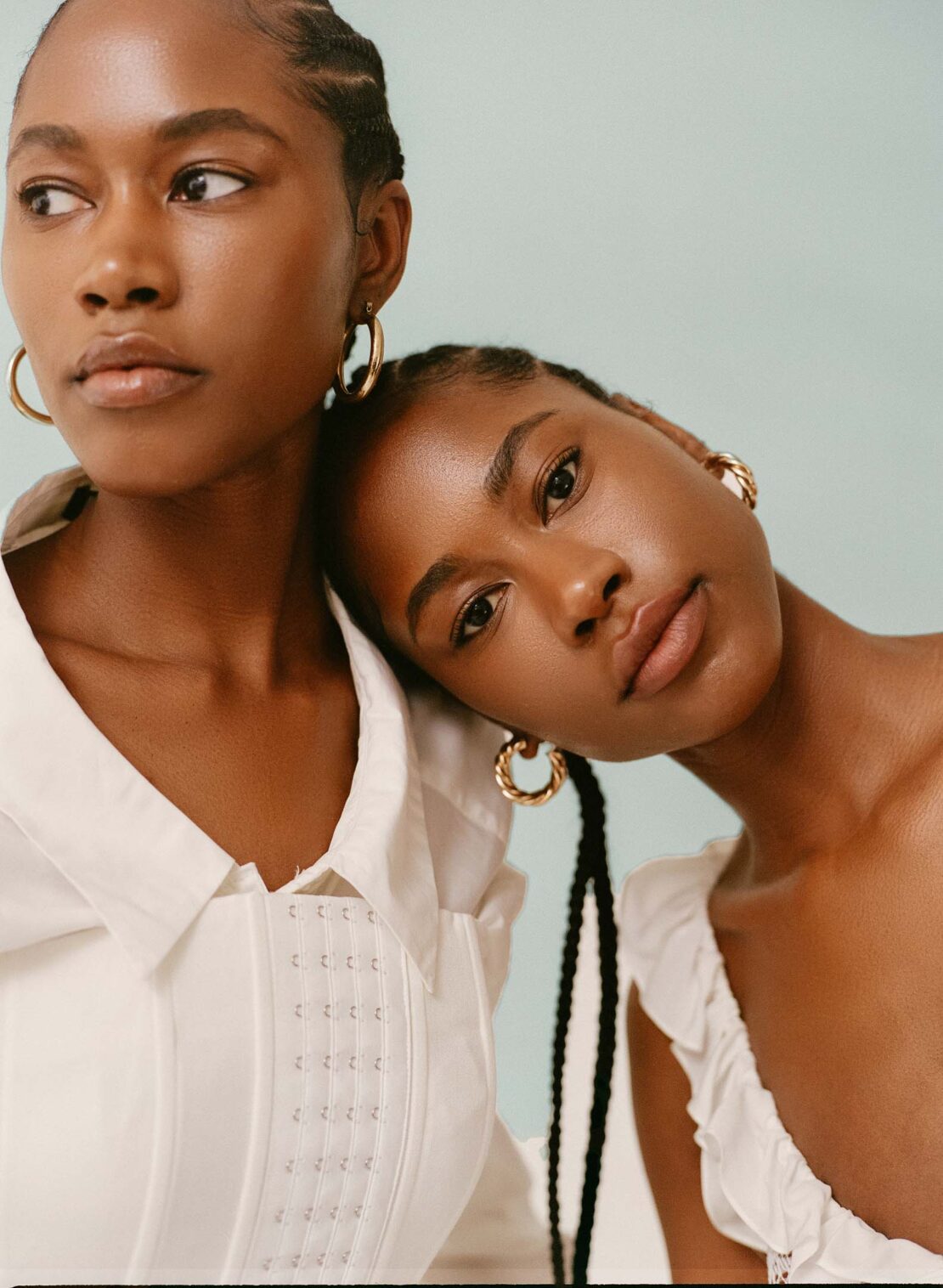 Two Black women in a close-up studio portrait. One rests her head on the shoulder of the other. Both are wearing white tops and gold earrings.