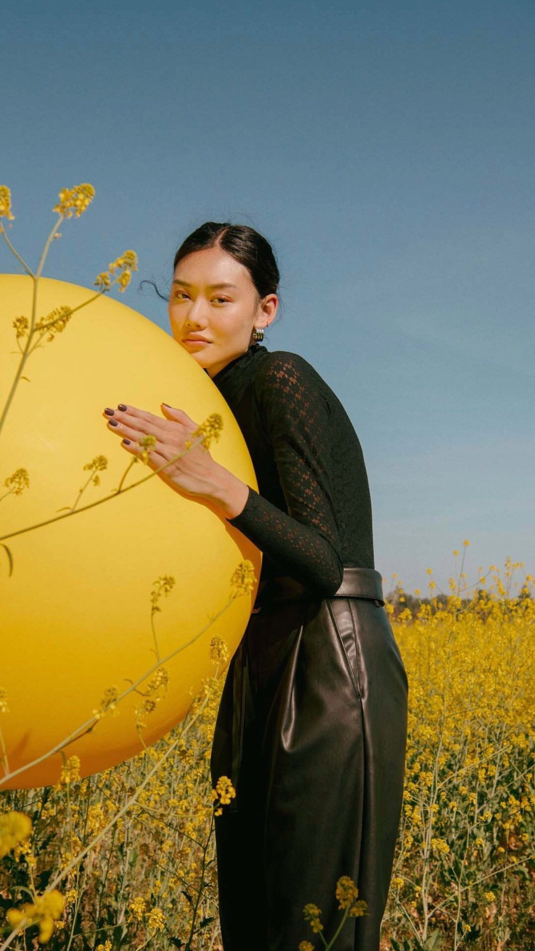 An Asian woman poses in a field of yellow flowers, holding a large yellow balloon. She wears a black top and leather pants against a blue sky.