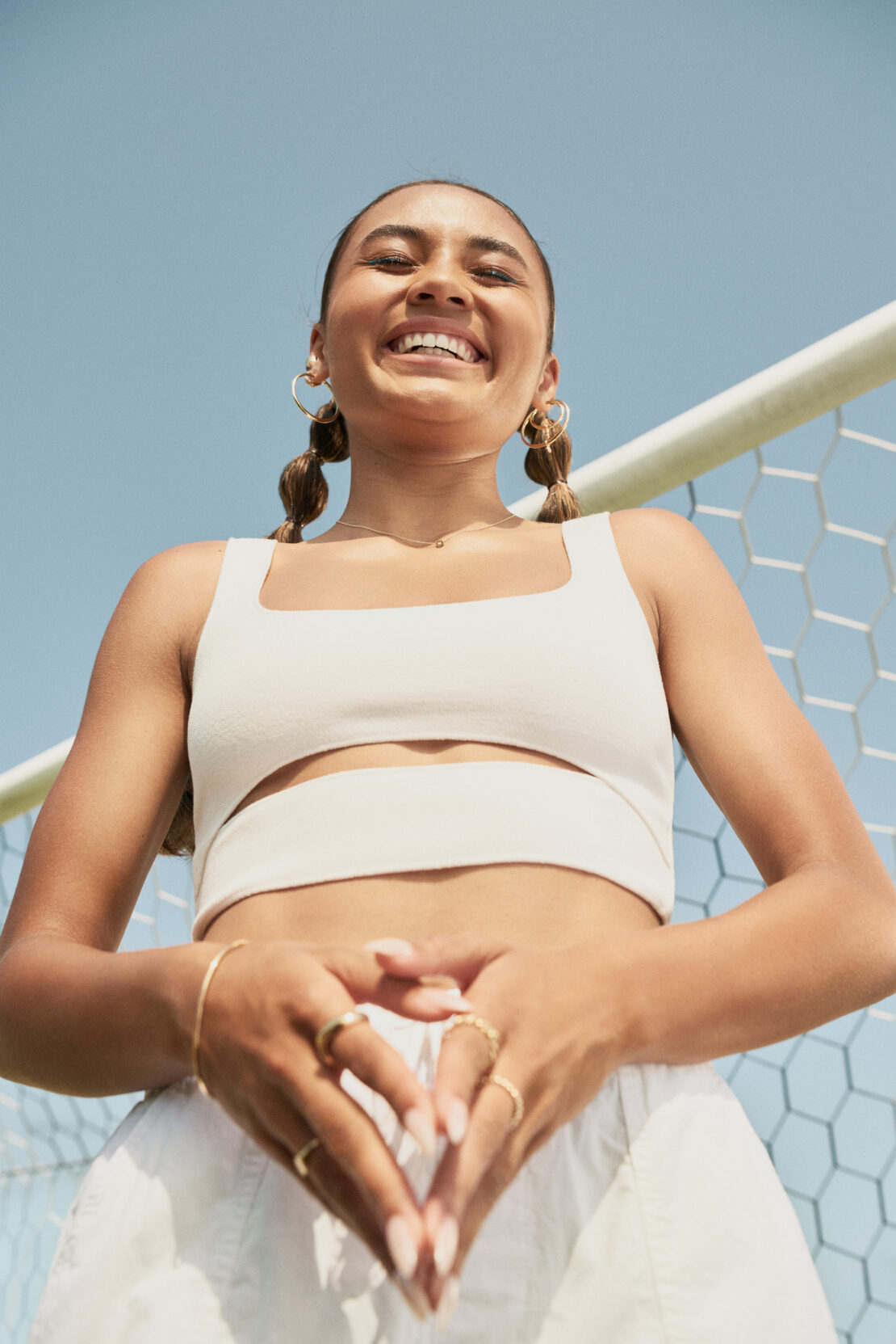 Smiling woman poses in front of soccer net, wearing a white crop top and shorts. She has her hands together and wears gold jewelry.