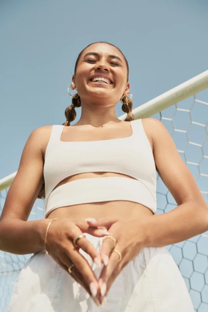 Smiling woman poses in front of soccer net, wearing a white crop top and shorts. She has her hands together and wears gold jewelry.