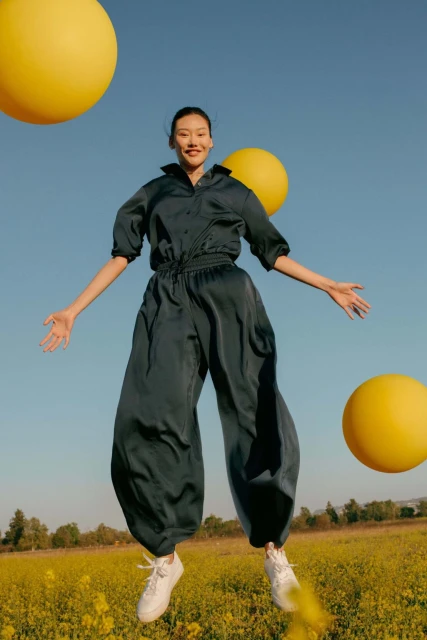 A young woman in a blue jumpsuit is jumping in a field of yellow flowers with yellow balloons against a bright blue sky.
