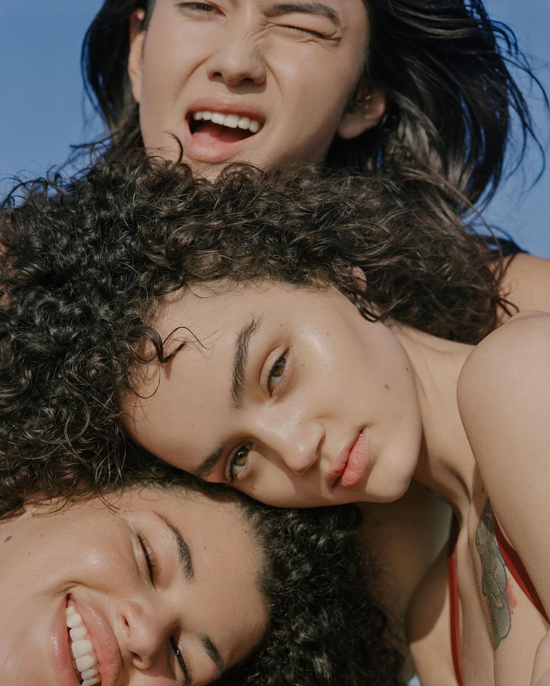 Three women pose closely together against a light blue sky. The women have diverse features and curly hair, and are smiling or winking.