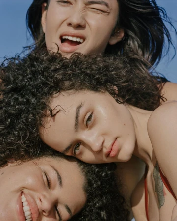 Three women pose closely together against a light blue sky. The women have diverse features and curly hair, and are smiling or winking.