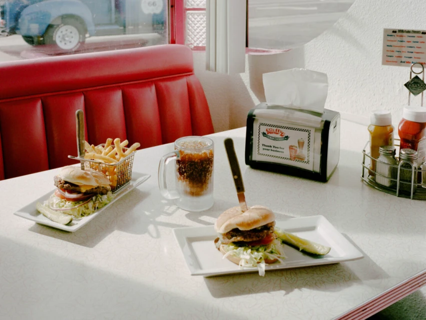 Diner food still life: cheeseburgers with fries, cola, and condiments on a white table with a red booth, in front of a window.