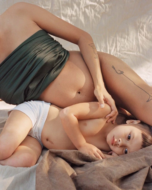 A pregnant woman with a green wrap poses with a young child in white shorts on a neutral-toned blanket indoors, with soft lighting.