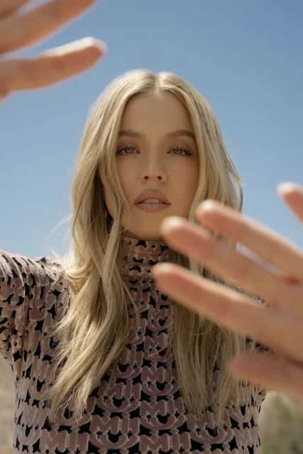 Portrait of a fair-skinned woman with blond hair, wearing a patterned mock turtleneck top, posing outdoors against a clear blue sky.