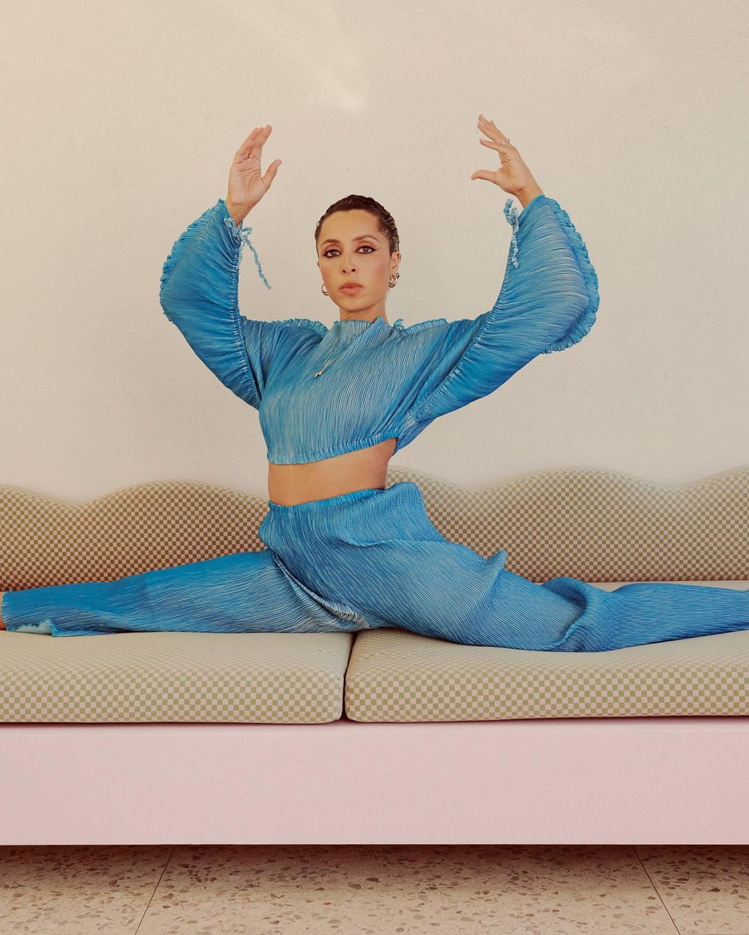 Adult female in a full split on a pink sofa. She's wearing matching aqua pleated pants and a crop top in front of a neutral wall.