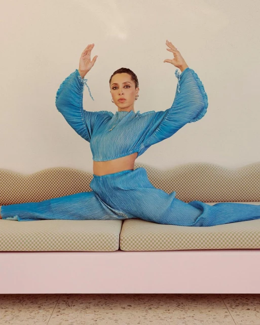 Adult female in a full split on a pink sofa. She's wearing matching aqua pleated pants and a crop top in front of a neutral wall.