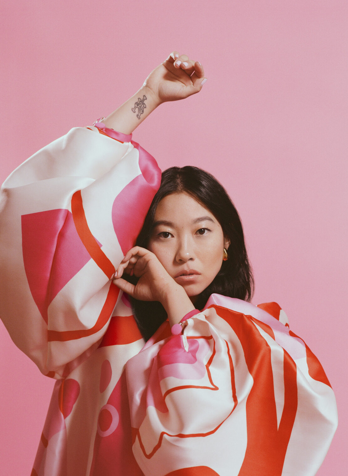 Portrait of an Asian woman in a patterned dress against a solid pink backdrop, posing with hands raised, captured in studio lighting.