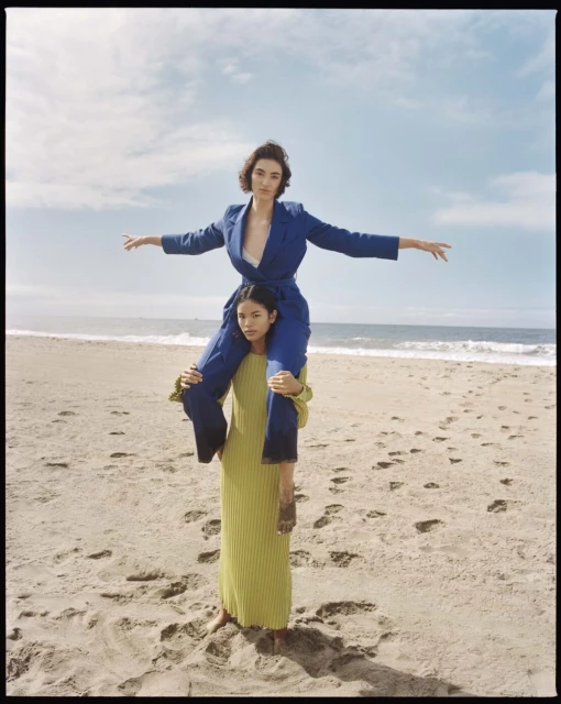 Two women pose on a beach with one woman on the shoulders of the other, wearing stylish clothing in a medium shot with the ocean.