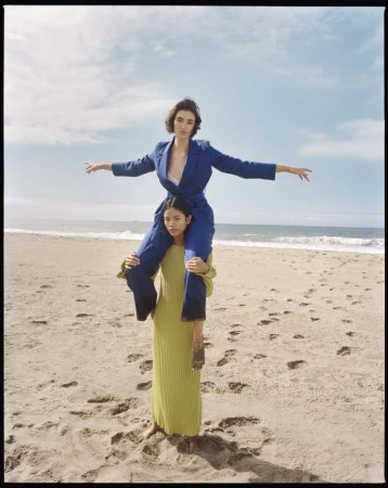 Two women pose on a beach with one woman on the shoulders of the other, wearing stylish clothing in a medium shot with the ocean.