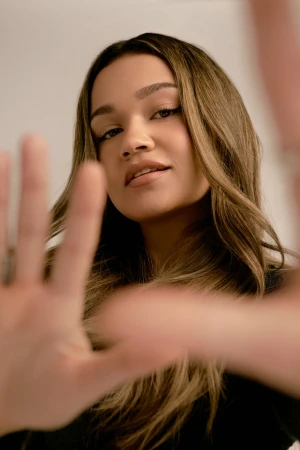 Close-up portrait of a woman with long highlighted hair reaching towards the camera with out-of-focus hands in the foreground.