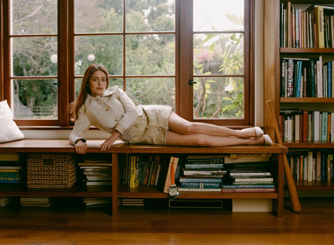 Adult woman with auburn hair poses in a beige jacket and skirt while reclining on a window seat with books and natural lighting.