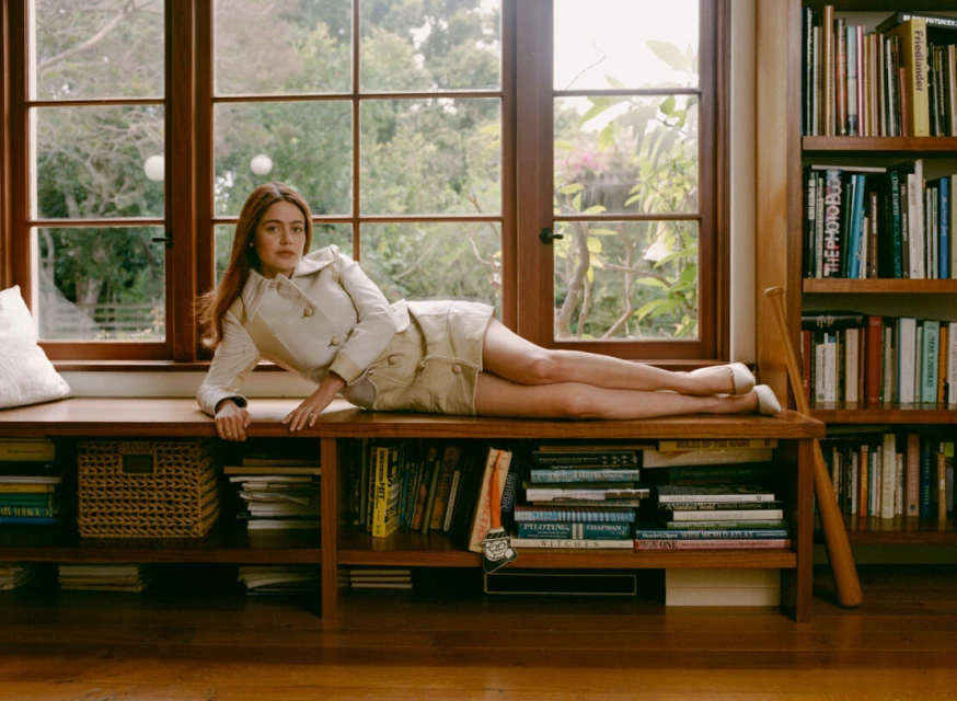 Adult woman with auburn hair poses in a beige jacket and skirt while reclining on a window seat with books and natural lighting.