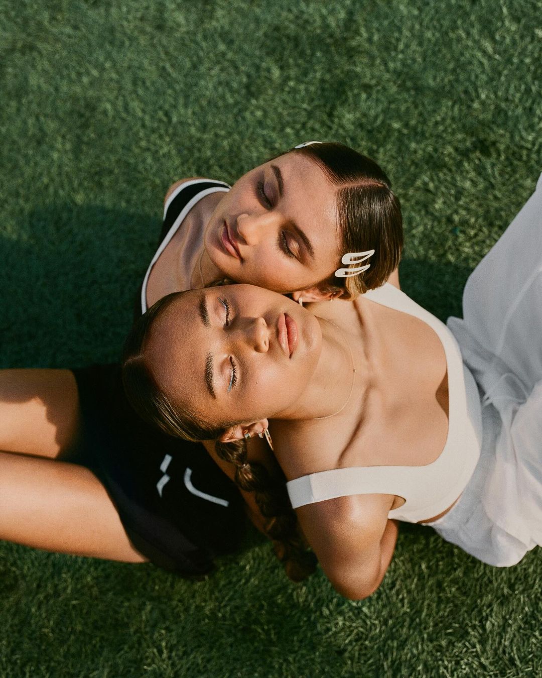 Two women lie on green grass in an overhead shot. One woman wears white activewear; the other wears black activewear. Both have closed eyes.