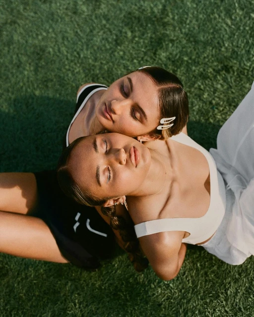 Two women lie on green grass in an overhead shot. One woman wears white activewear; the other wears black activewear. Both have closed eyes.