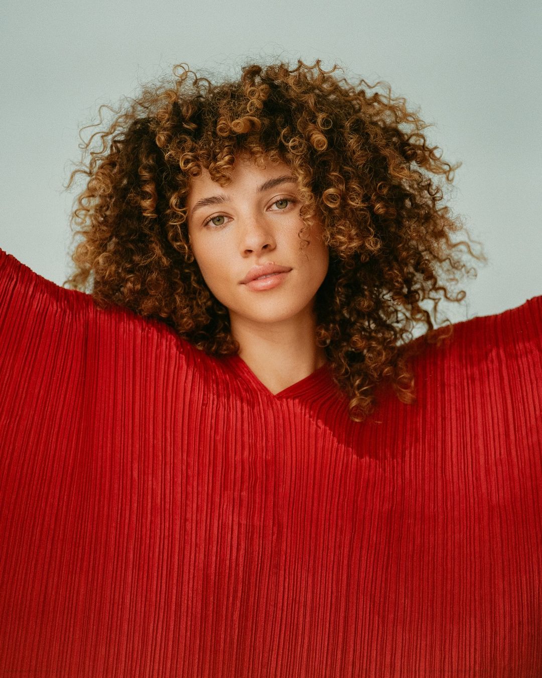 Portrait of a woman with curly brown hair wearing a flowing, textured red top, captured with soft studio lighting and a light blue background.