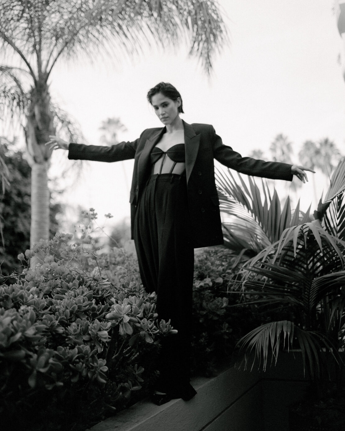 A woman in a dark bra top and jacket stands among tropical plants and trees in a black-and-white fashion shot, with arms outstretched.