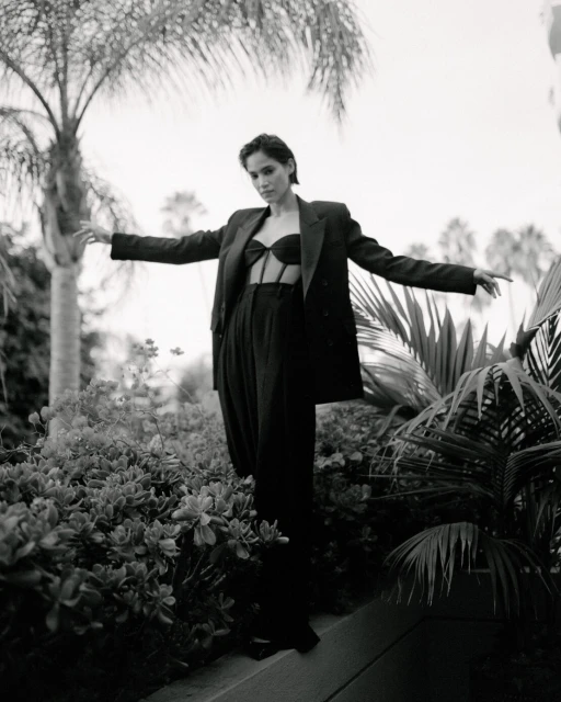 A woman in a dark bra top and jacket stands among tropical plants and trees in a black-and-white fashion shot, with arms outstretched.