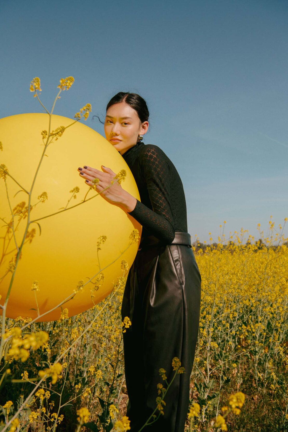 A woman poses with a large yellow balloon in a field of yellow wildflowers under a bright blue sky in an outdoor fashion shot.