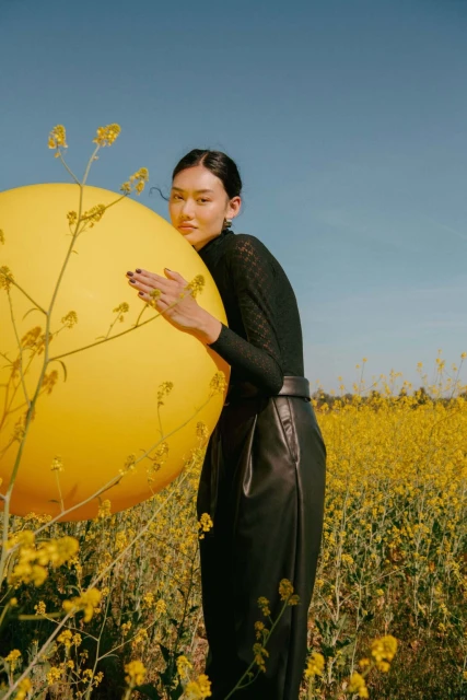 A woman poses with a large yellow balloon in a field of yellow wildflowers under a bright blue sky in an outdoor fashion shot.