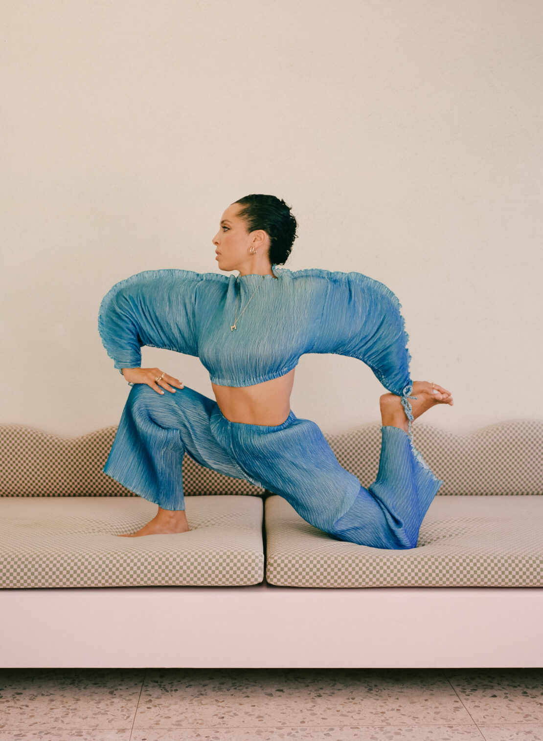 Woman in blue textured crop top and wide-leg pants poses barefoot on a cream-colored couch against a plain cream wall in a studio.
