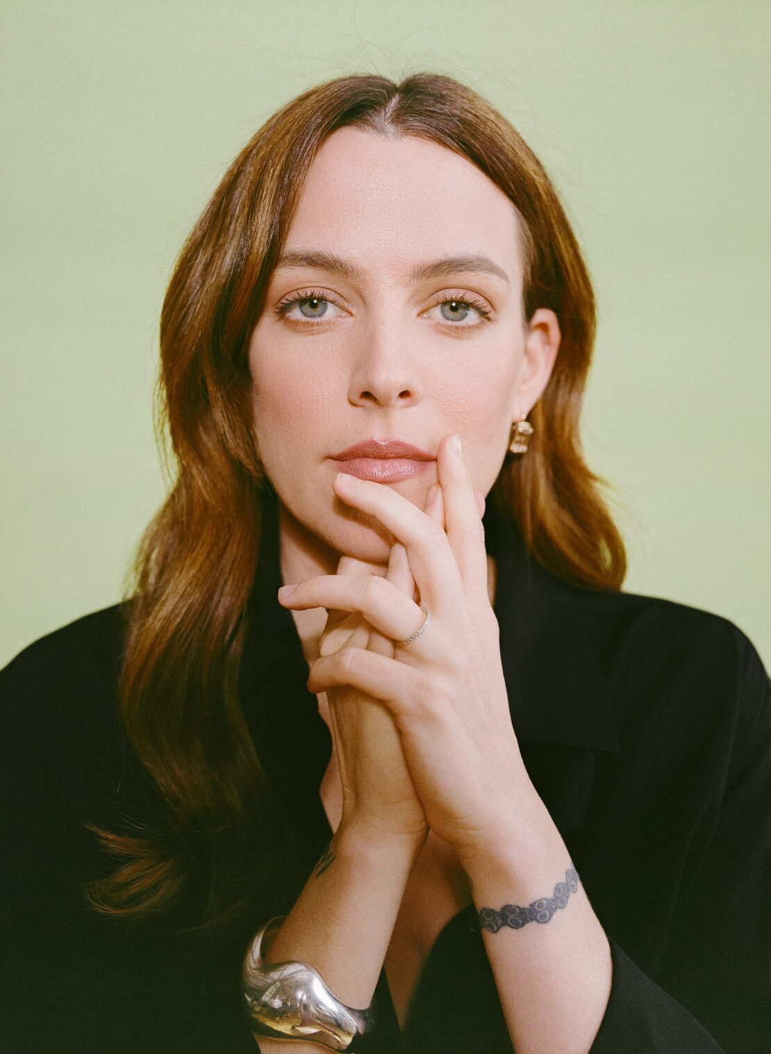 Portrait of a woman with brown hair holding her hands up near her face, wearing a black top and silver jewelry, against a green background.