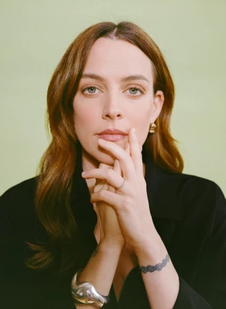 Portrait of a woman with brown hair holding her hands up near her face, wearing a black top and silver jewelry, against a green background.