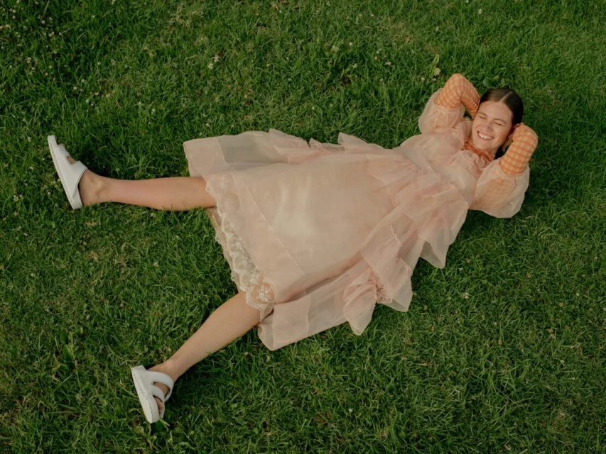 A woman with brown hair is lying on grass in a pink dress and white sandals, smiling with her hands behind her head.