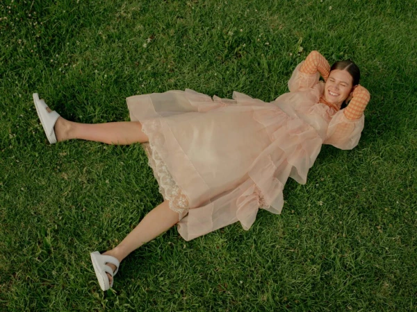 A woman with brown hair is lying on grass in a pink dress and white sandals, smiling with her hands behind her head.