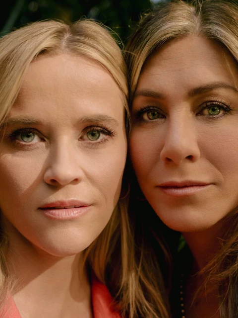 Close-up portrait of actresses Reese Witherspoon and Jennifer Aniston, side-by-side, showcasing their similar hair and eye color outdoors.