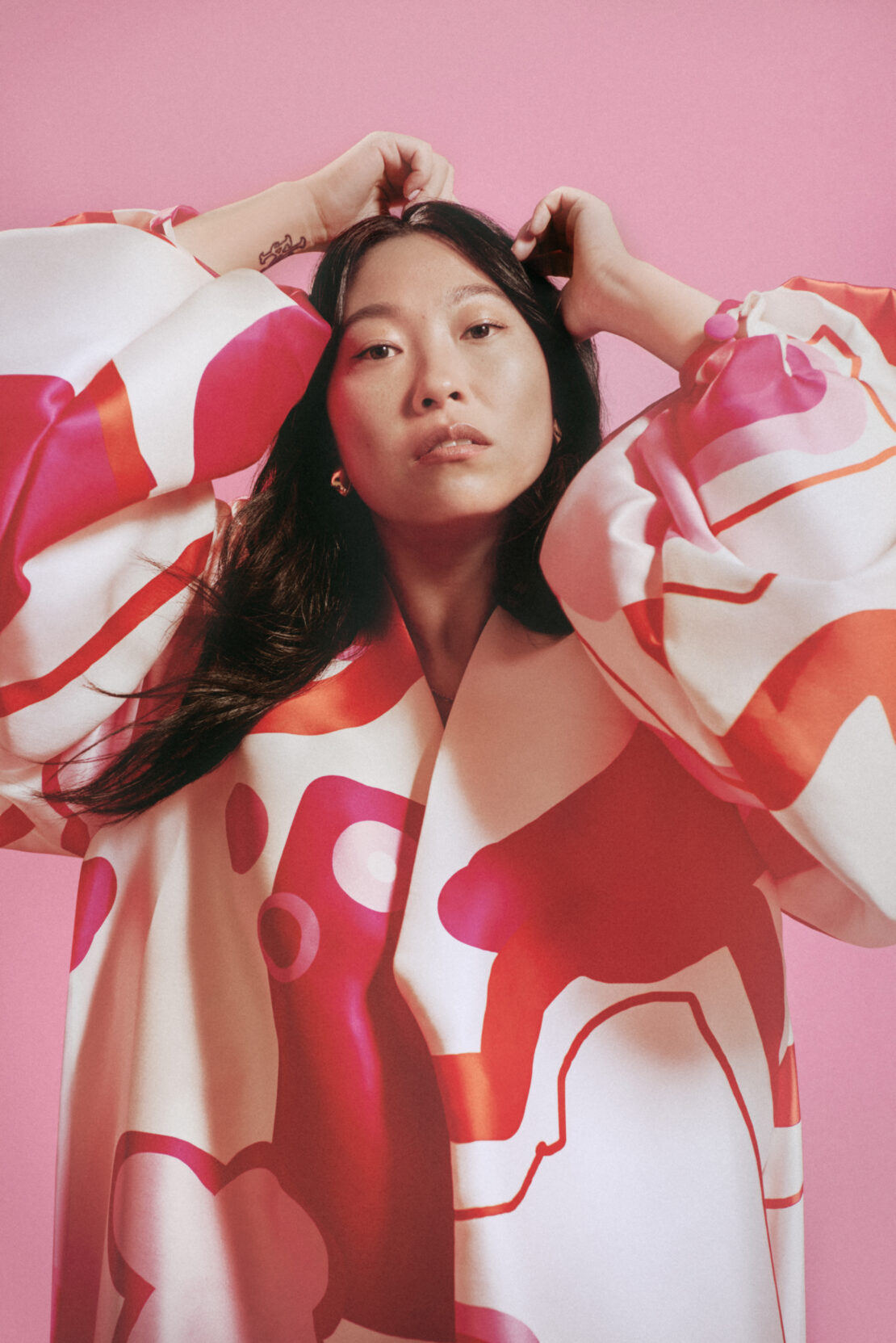 Portrait of an Asian woman in a patterned white and coral robe, with her hands near her head and a pink background.