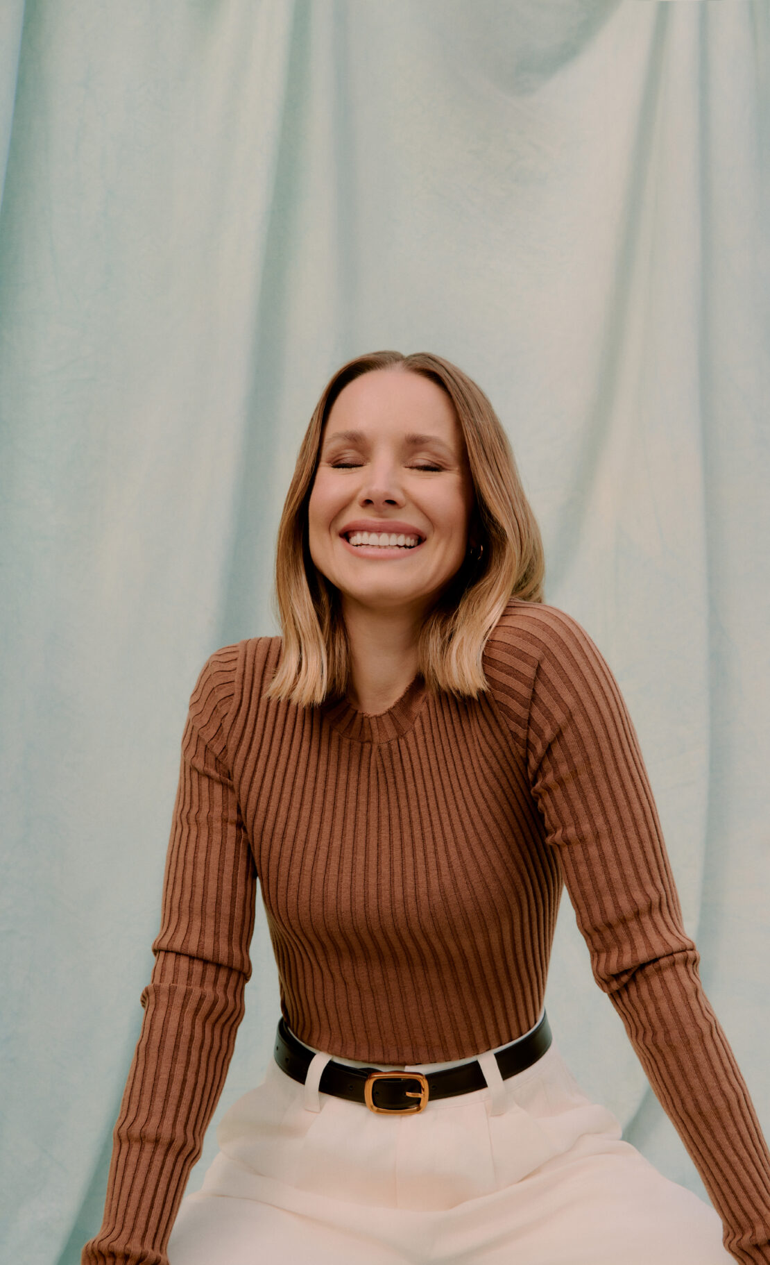 Close-up studio shot of smiling adult woman with blonde hair wearing a ribbed sweater and cream-colored slacks with a black belt.