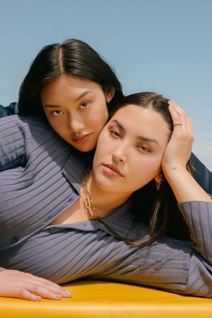 Two women in light blue tops pose closely on a yellow surface under a bright blue sky. They both wear necklaces and earrings.