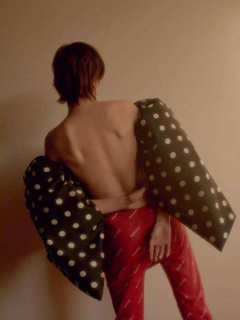 Person from the back wearing red pants and black polka dot arm coverings against a beige wall in a studio setting.