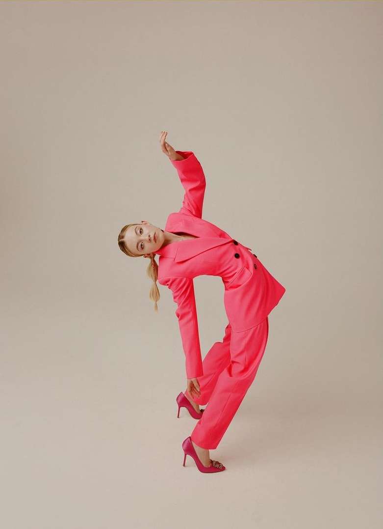 Elegant woman in a vibrant pink pantsuit strikes a pose against a neutral background, showcasing fashion and style with a dynamic posture.