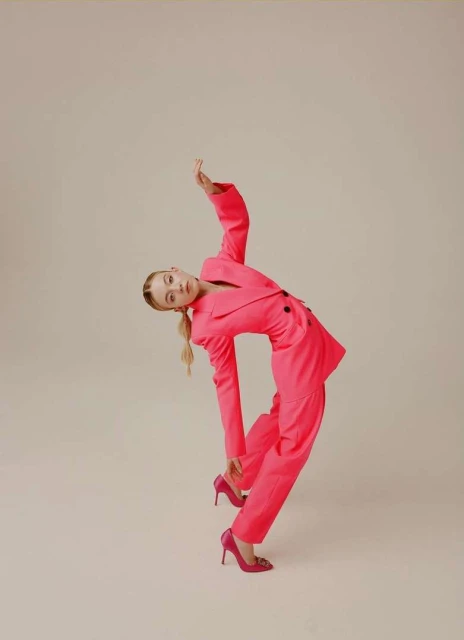 Elegant woman in a vibrant pink pantsuit strikes a pose against a neutral background, showcasing fashion and style with a dynamic posture.