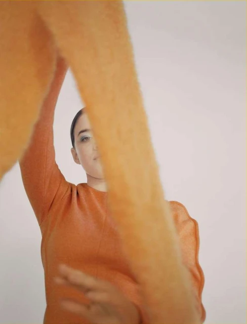 Woman in an orange sweater poses against a plain background. The fuzzy orange sleeve is dominant in the foreground with her facial features visible.