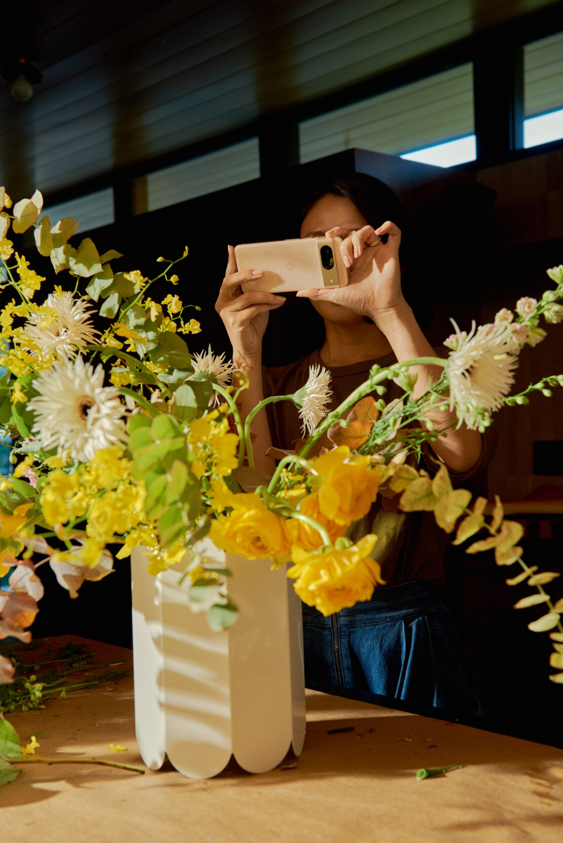 A woman holds up a smartphone to photograph a vibrant floral arrangement in a white vase on a table indoors, filtered sunlight.