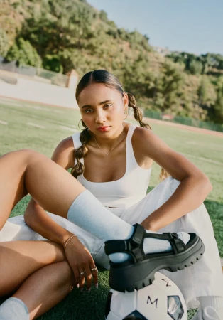 A woman with pigtails poses on a soccer field with a soccer ball, wearing a white top, light-colored pants, and platform sandals.
