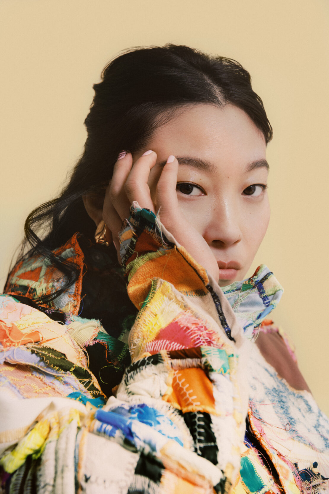Portrait of an Asian female model in a colorful patchwork jacket, hand near face, against a soft beige background.