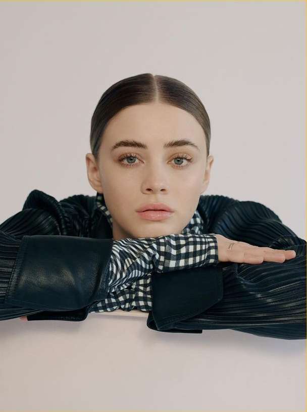 Close-up portrait of a young woman with sleek hair, wearing a black and white patterned blouse and black jacket, posing in a photo studio.