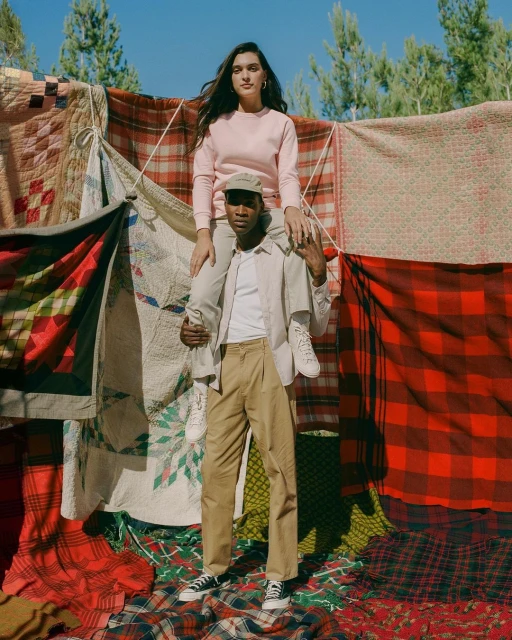 Woman sits on a man's shoulders amidst a colorful patchwork and plaid fabric backdrop in an outdoor setting with tree background.