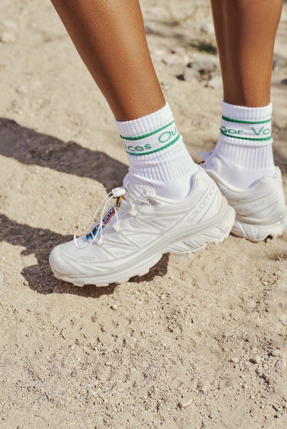 Close-up of a woman wearing white Salomon running shoes and white socks with green stripes standing on light brown dirt.