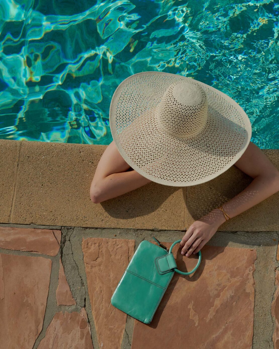 Woman in a wide-brimmed hat relaxing by the pool with a turquoise handbag. Summer leisure, vacations, poolside fashion.
