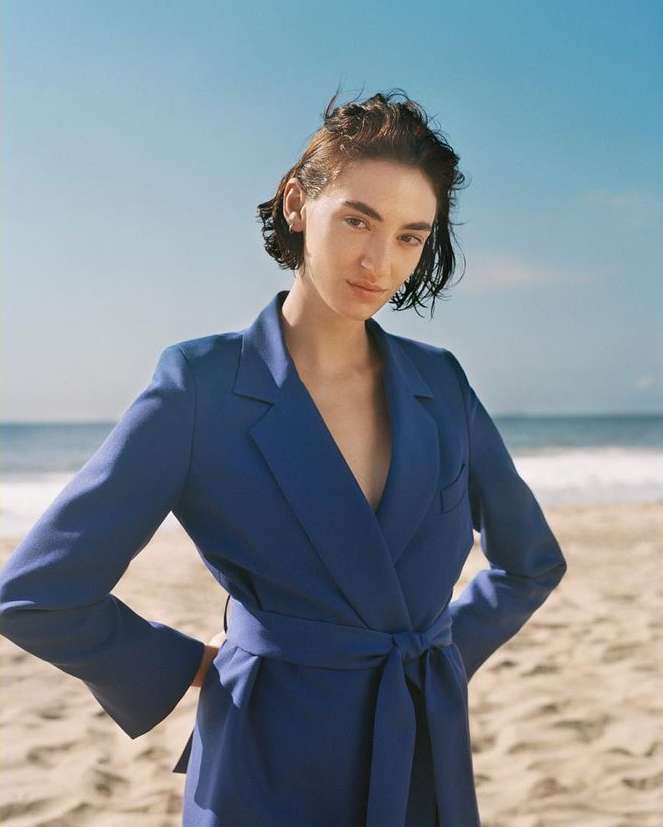 Model wearing a blue blazer on a sandy beach with ocean waves in the background.