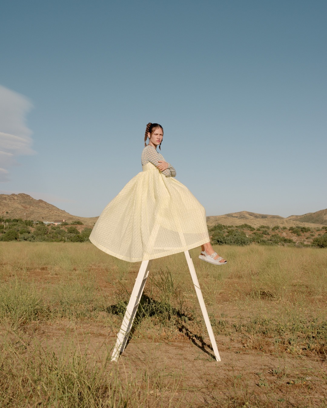 A young woman sits atop a white wooden sawhorse in a meadow under a clear blue sky, wearing a polka-dot yellow dress.