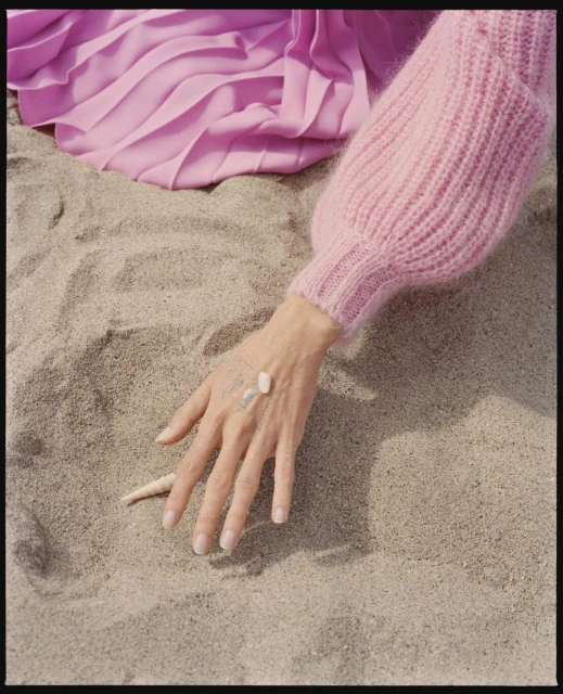 Hand with shells on sand in light pink sweater and skirt; natural light, soft textures, summer aesthetic, beach vibe, editorial.
