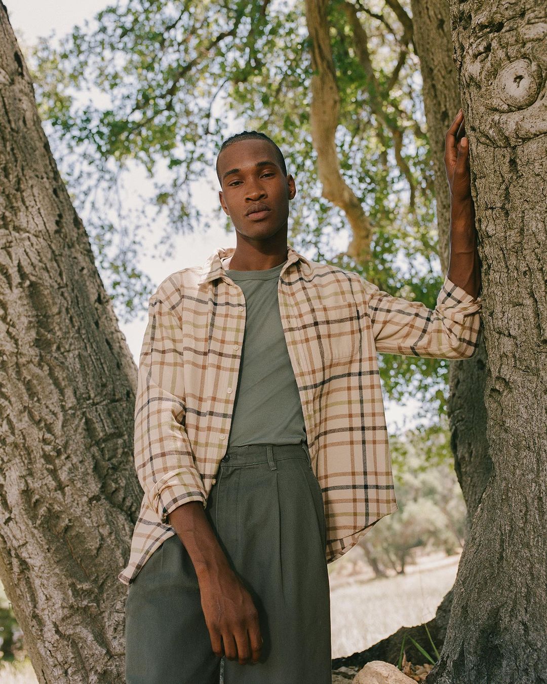 A black man wearing a flannel shirt and green pants poses next to a tree in an outdoor setting with natural lighting and lush background.