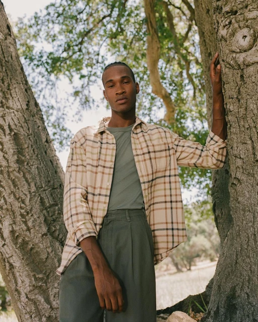 A black man wearing a flannel shirt and green pants poses next to a tree in an outdoor setting with natural lighting and lush background.